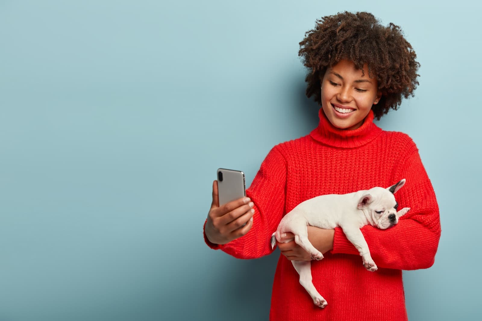 A smiling woman in a red knitted sweater takes a selfie on a phone while holding a sleeping French bulldog puppy against a plain blue background.