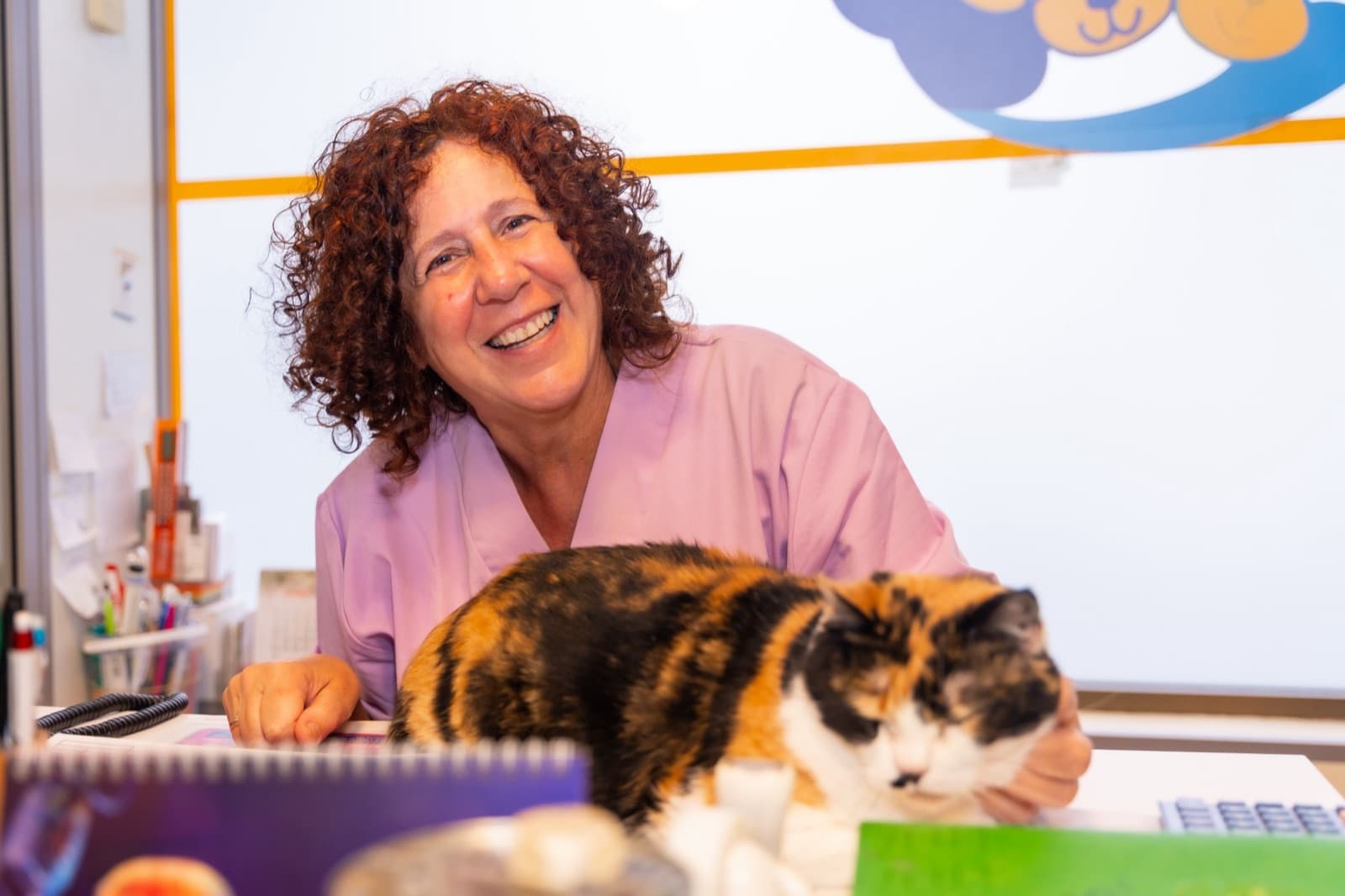 A smiling staff member in a pink scrub top leans over a veterinary clinic front desk while a calico cat dozes beside the phone.
