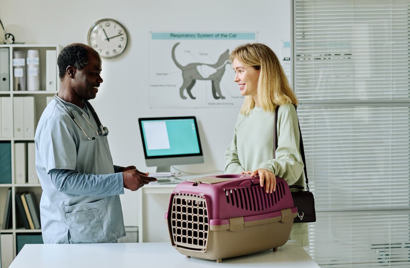A veterinarian in light blue scrubs holds a clipboard while talking with a smiling client who rests both hands on a plum pet carrier in an exam room.
