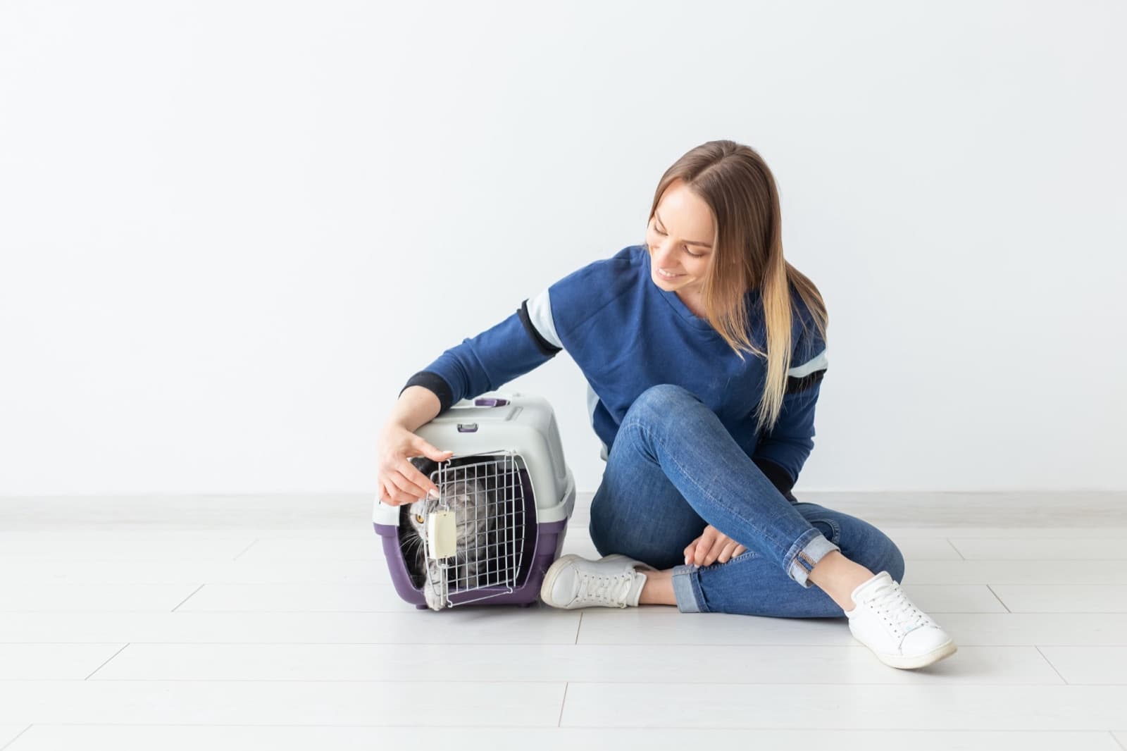 A woman in a blue sweatshirt and jeans sits on a white floor beside a purple and grey pet carrier, reaching in to reassure the grey cat inside.