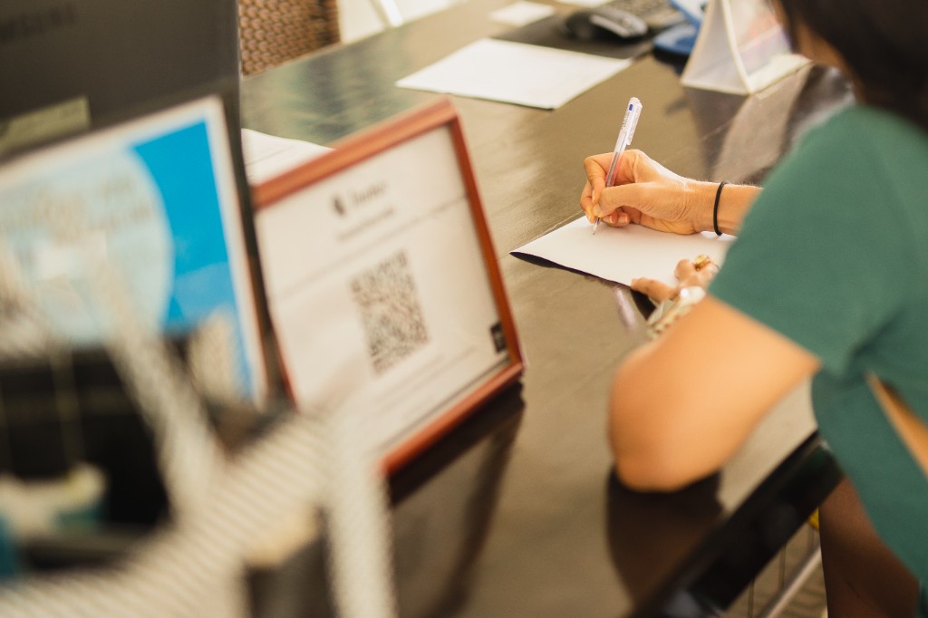 A staff member completes paperwork with a blue pen at a dark front desk, a QR code sign blurred in the foreground.